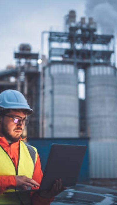 A shot of an young engineer wearing a helmet and using a laptop and hands free device during his night shirt in the oil rafinery. Engineering concept.